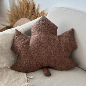 May include: A brown leaf-shaped decorative pillow with a textured, plush surface. The pillow is resting on a light-colored sofa. The pillow's design resembles a maple leaf, adding a touch of nature-inspired decor to any living space.
