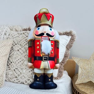 Puede incluir: Una almohada de cascanueces roja y dorada con barba blanca y sombrero negro. La almohada está sentada en un sofá blanco con una almohada de tiro de color crema.