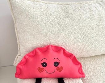 May include: A plush, pink dumpling-shaped pillow with a smiling face, button eyes, and heart-shaped cheeks. It has black legs and is resting on a white, textured surface, likely a couch or bed.