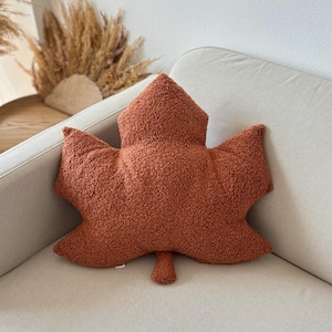 Könnte beinhalten: Ein kissenförmiges Dekokissen in Rostbraun, geformt wie ein Blatt mit strukturierter Oberfläche. Das Kissen ist in Form eines Ahornblattes mit ausgeprägten Lappen und einem Stiel gestaltet. Das Kissen liegt auf einem neutralfarbenen Sofa.