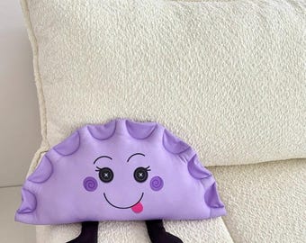May include: A purple plush pillow shaped like a half-moon with a smiling face, two eyes, and a tongue sticking out. The pillow has two black legs.
