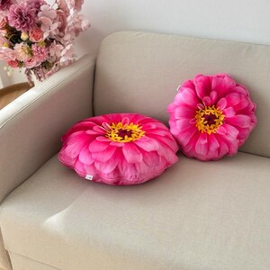 May include: Two pink flower-shaped throw pillows with yellow centers on a white couch.