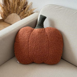 May include: A textured, orange pumpkin-shaped cushion with a green stalk. The cushion is placed on a beige sofa, evoking an autumnal atmosphere. The cushion is a decorative item, perfect for the autumn season.