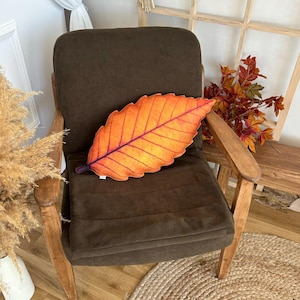 May include: A brown armchair with a large orange leaf-shaped pillow. The pillow is made of fabric and has a realistic leaf design.