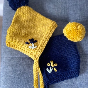 Könnte beinhalten: Zwei gestrickte Babyhauben. Eine ist senfgelb mit einem marineblauen Bommel und einer gestickten Blume. Die andere ist marineblau mit einem gelben Bommel und einer gestickten Blume. Beide Hauben haben gelbe Bänder.
