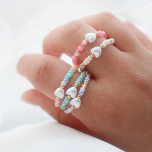 Könnte beinhalten: Vier farbige Perlenringe mit silbernen Herzanhängern. Die Ringe sind auf einem Finger übereinander gelegt.