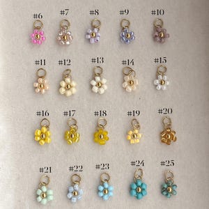 May include: A collection of 27 flower-shaped charms in various colors, each with a gold-colored loop. The charms are made of small beads and are arranged in rows. The colors include pink, yellow, blue, and green.