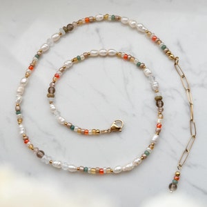 Könnte beinhalten: Eine zarte goldene Kettenkette mit einem kleinen Anhänger am Ende. Die Kette besteht aus kleinen weißen Perlen, orangefarbenen, grünen und braunen Perlen.