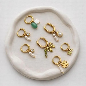 Può includere: Una collezione di sette orecchini a cerchio dorati con vari ciondoli, tra cui una pietra verde, perline bianche, un piccolo fiocco, una lettera S, un ciondolo a forma di fiore e un pendente circolare testurizzato, disposti su un piatto in ceramica.