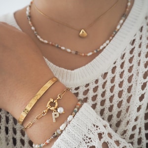 May include: A gold chain necklace with a small shell pendant and a multi-colored beaded necklace. The person is also wearing three gold bracelets, one with a charm that has a pearl and a letter "P" on it.