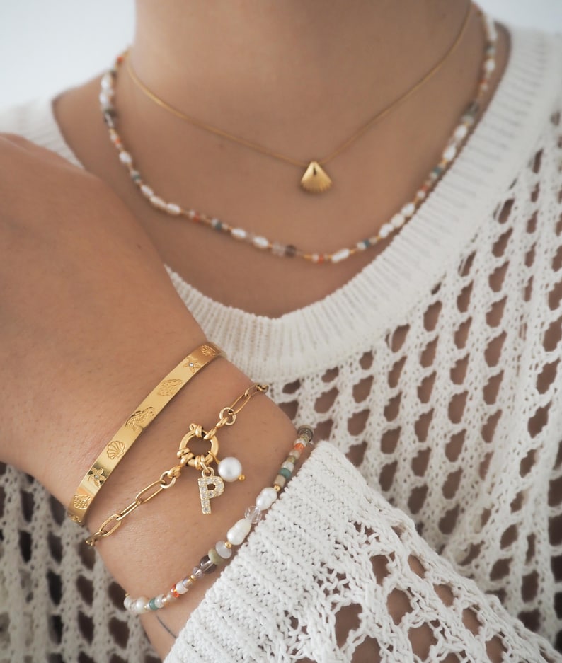 May include: A gold chain necklace with a small shell pendant and a multi-colored beaded necklace. The person is also wearing three gold bracelets, one with a charm that has a pearl and a letter "P" on it.
