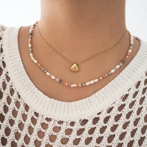 May include: A layered necklace with a gold shell pendant. The top necklace is a delicate gold chain. The bottom necklace is made of small white pearls, peach, and brown beads.