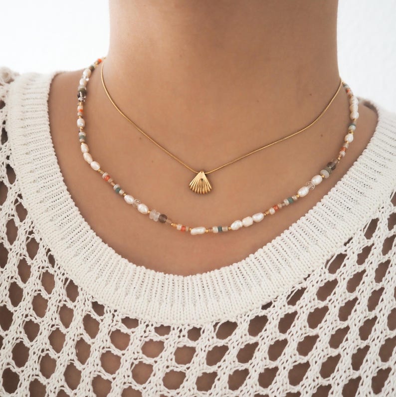 May include: A layered necklace with a gold shell pendant. The top necklace is a delicate gold chain. The bottom necklace is made of small white pearls, peach, and brown beads.