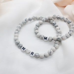 May include: Two white marble beaded bracelets with black letter charms. The larger bracelet has the letter "M" and the smaller bracelet has the letters "N", a heart charm, and "R".