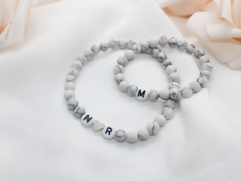 May include: Two white marble beaded bracelets with black letter charms. The larger bracelet has the letter "M" and the smaller bracelet has the letters "N", a heart charm, and "R".