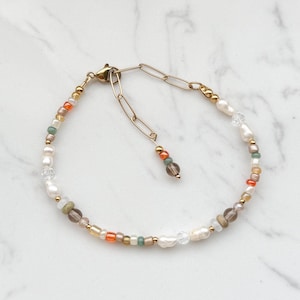 May include: A delicate gold chain bracelet with a variety of small beads in shades of white, cream, orange, brown, and green. The bracelet features a lobster clasp closure.