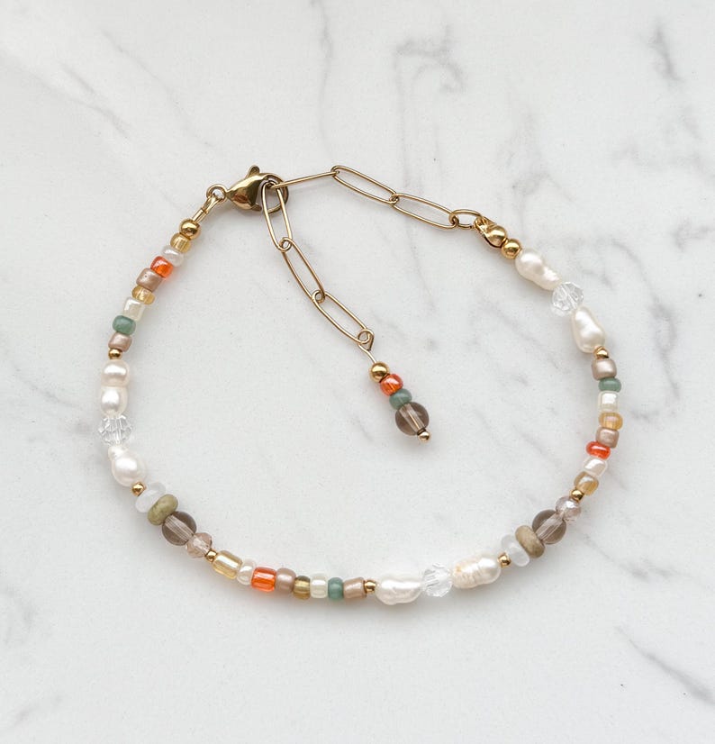 May include: A delicate gold chain bracelet with a variety of small beads in shades of white, cream, orange, brown, and green. The bracelet features a lobster clasp closure.