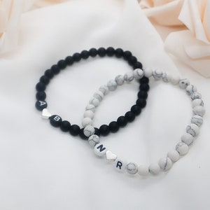 May include: Two beaded bracelets, one black and one white with gray marbling. The black bracelet has the letters "AB" with a silver heart charm. The white bracelet has the letters "NR" with a silver heart charm.