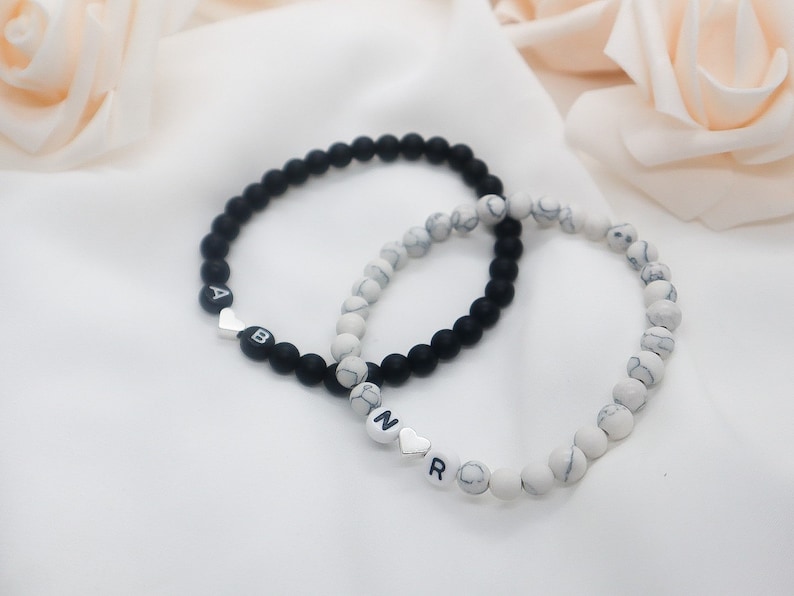 May include: Two beaded bracelets, one black and one white with gray marbling. The black bracelet has the letters "AB" with a silver heart charm. The white bracelet has the letters "NR" with a silver heart charm.