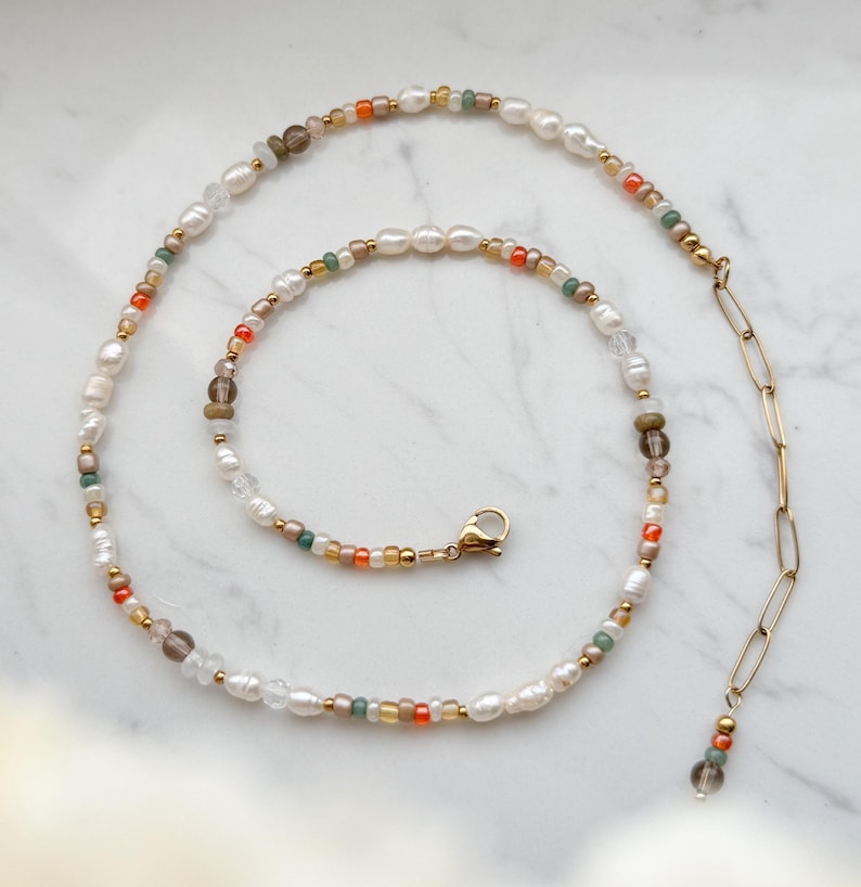 May include: A delicate gold chain necklace with a small charm at the end. The necklace is made of small white pearls, orange, green, and brown beads.