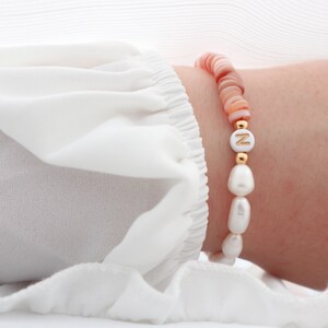 Puede incluir: Un brazalete con cuentas rosas y blancas, incluyendo una cuenta blanca con una letra dorada 'N'.