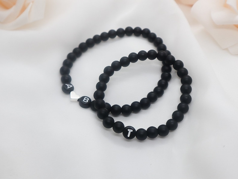 May include: Two black beaded bracelets with silver heart and letter charms. The bracelets spell out "AB" and "T".