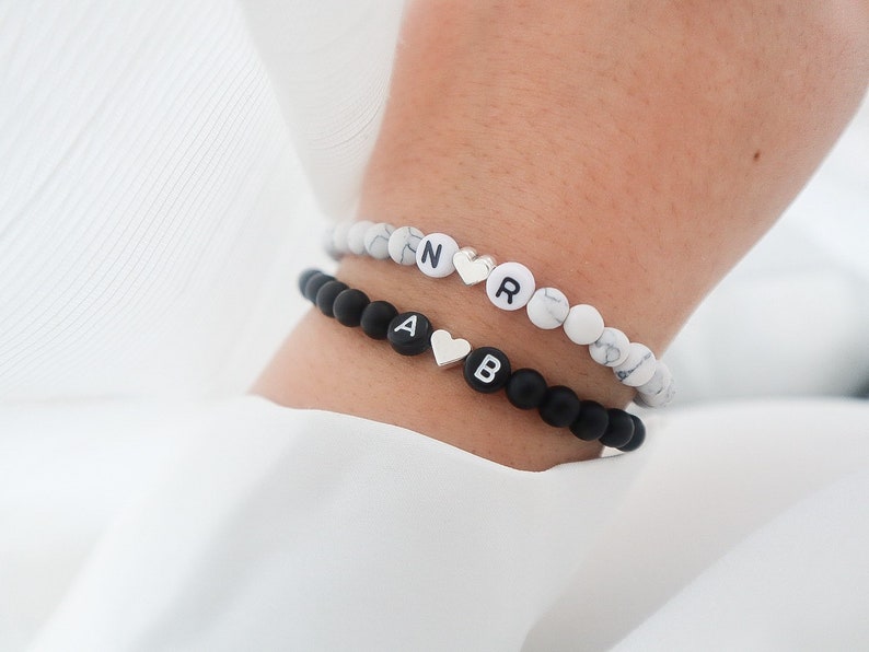 May include: Two beaded bracelets with silver heart charms. The top bracelet has white beads with black letters spelling "NR" and the bottom bracelet has black beads with white letters spelling "AB".