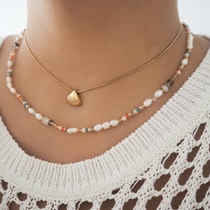 May include: A gold chain necklace with a small seashell pendant layered over a beaded necklace with white pearls, orange, brown, and green beads.