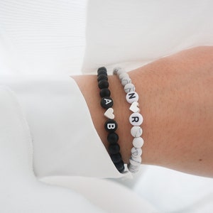 May include: Two beaded bracelets with black and white beads. One bracelet has black beads with white letters spelling "AB" and a silver heart charm. The other bracelet has white beads with black letters spelling "NR" and a silver heart charm.