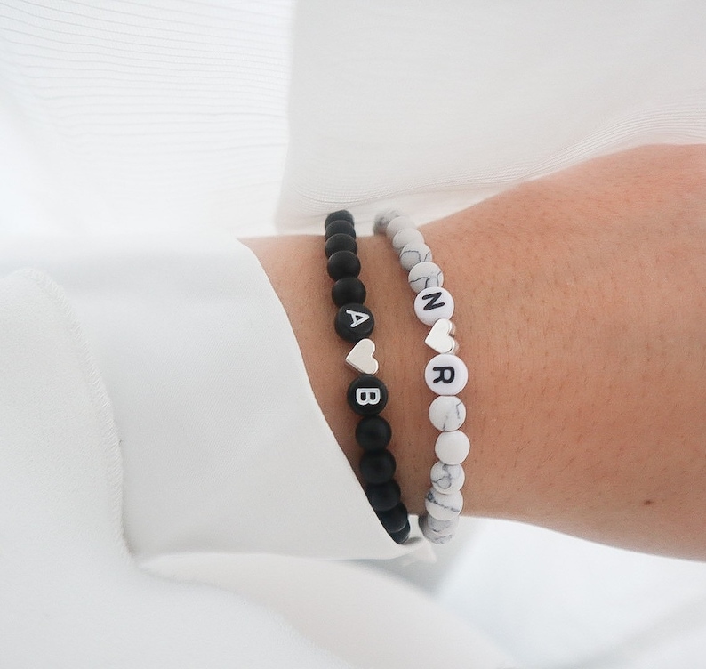 May include: Two beaded bracelets with black and white beads. One bracelet has black beads with white letters spelling "AB" and a silver heart charm. The other bracelet has white beads with black letters spelling "NR" and a silver heart charm.