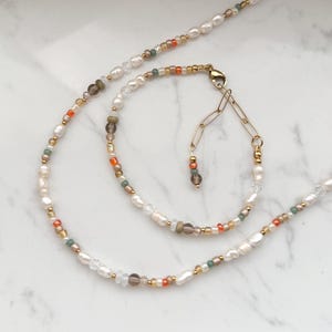 May include: A delicate necklace and bracelet set featuring white pearls, brown, orange, and green beads. The necklace and bracelet are connected by a gold chain.