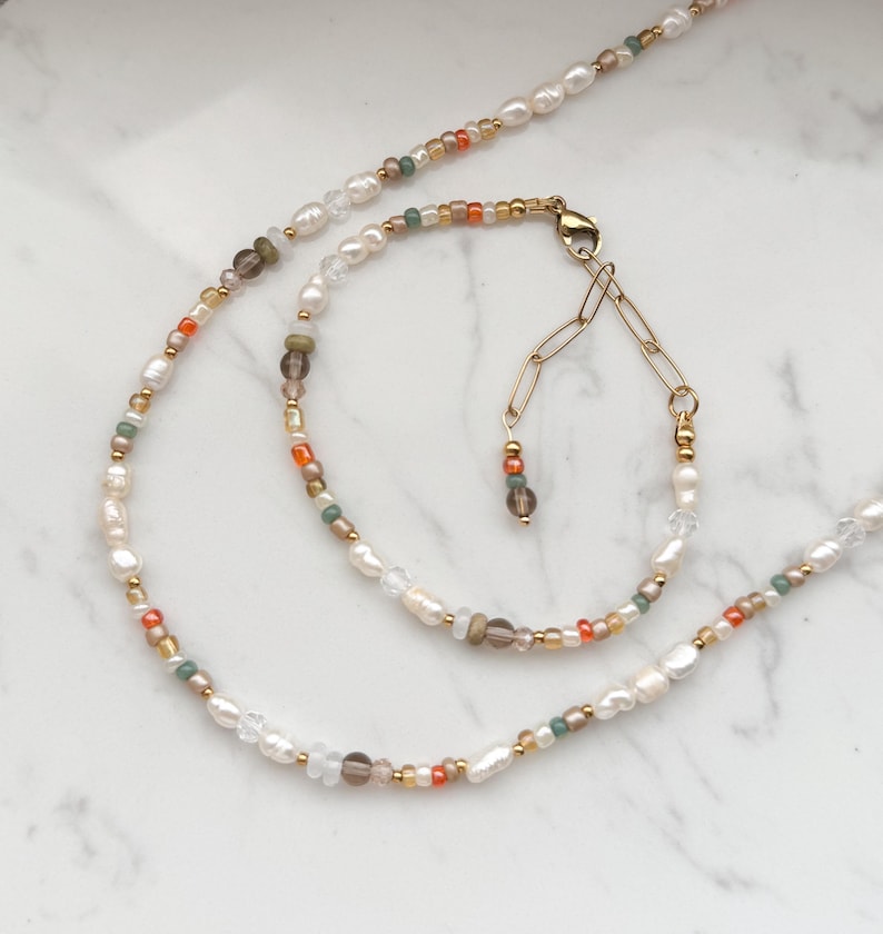 May include: A delicate necklace and bracelet set featuring white pearls, brown, orange, and green beads. The necklace and bracelet are connected by a gold chain.