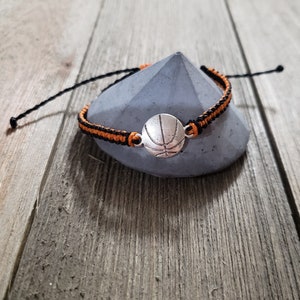 May include: A black and orange braided bracelet with a silver basketball charm.