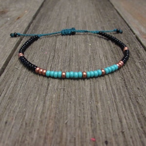 May include: A turquoise and black beaded bracelet with copper accents. The bracelet is tied with a dark blue cord.