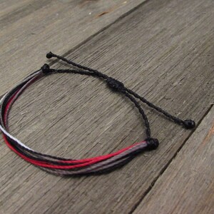 May include: Adjustable bracelet featuring a combination of red, black, gray, and white threads. The bracelet is made of multiple strands and has a sliding knot closure for a customizable fit. The bracelet is shown on a wooden surface.