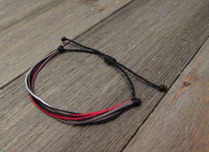 May include: Adjustable bracelet featuring a combination of red, black, gray, and white threads. The bracelet is made of multiple strands and has a sliding knot closure for a customizable fit. The bracelet is shown on a wooden surface.