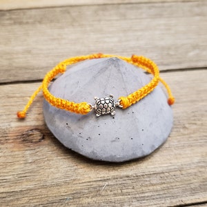 May include: A yellow friendship bracelet with a silver turtle charm. The bracelet is tied around a gray stone.