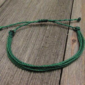 May include: A handmade green string bracelet. The bracelet is made of multiple strands of green string, knotted together to form a circular shape. The adjustable closure is also green. The bracelet is displayed on a weathered wooden surface.