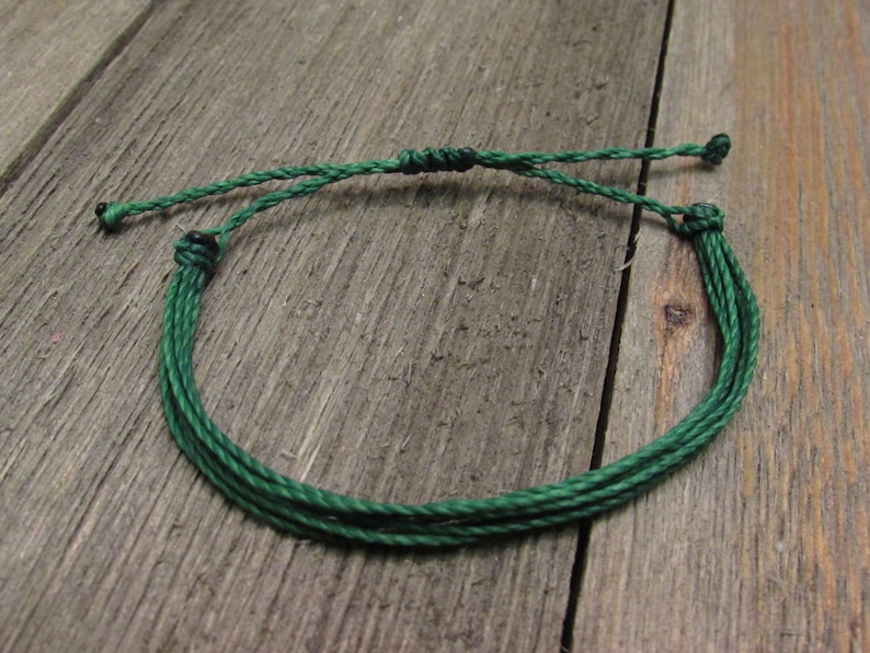 May include: A handmade green string bracelet. The bracelet is made of multiple strands of green string, knotted together to form a circular shape. The adjustable closure is also green. The bracelet is displayed on a weathered wooden surface.