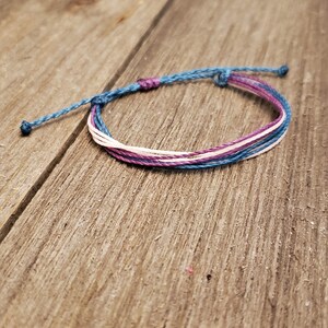 May include: A multi-colored friendship bracelet with blue, pink, and white threads. The bracelet is tied with a knot at the end.