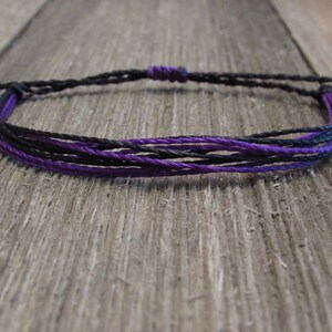 May include: A black and purple braided friendship bracelet. The bracelet is made of multiple strands of cord and is tied with a knot at the end.
