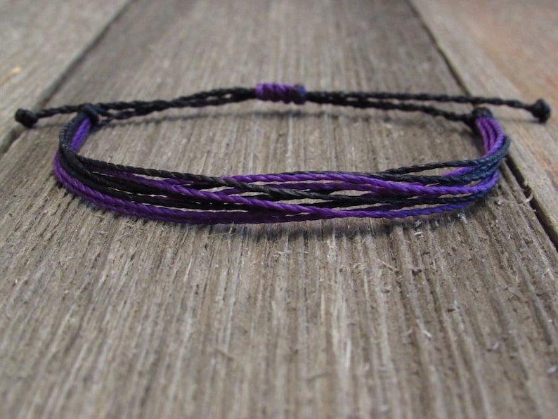 May include: A black and purple braided friendship bracelet. The bracelet is made of multiple strands of cord and is tied with a knot at the end.