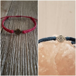 May include: Two bracelets with metal charms. The first bracelet is red and has a round charm. The second bracelet is blue and has a compass charm with the letters N, W, E, and S.