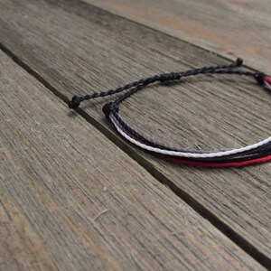 May include: A handmade bracelet featuring four strands of cord in black, gray, white, and red. The bracelet is knotted at both ends and rests on a weathered wooden surface. The bracelet is a simple, minimalist design.