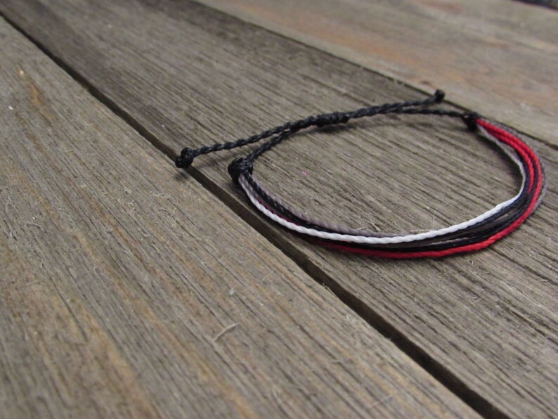 May include: A handmade bracelet featuring four strands of cord in black, gray, white, and red. The bracelet is knotted at both ends and rests on a weathered wooden surface. The bracelet is a simple, minimalist design.