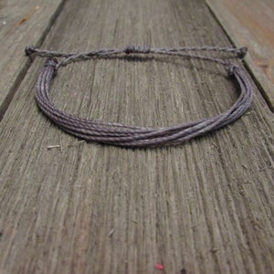 Op de afbeelding: Een grijs gevlochten armband gemaakt van meerdere strengen koord. De armband is vastgemaakt met een simpele knoop en ligt op een houten oppervlak.