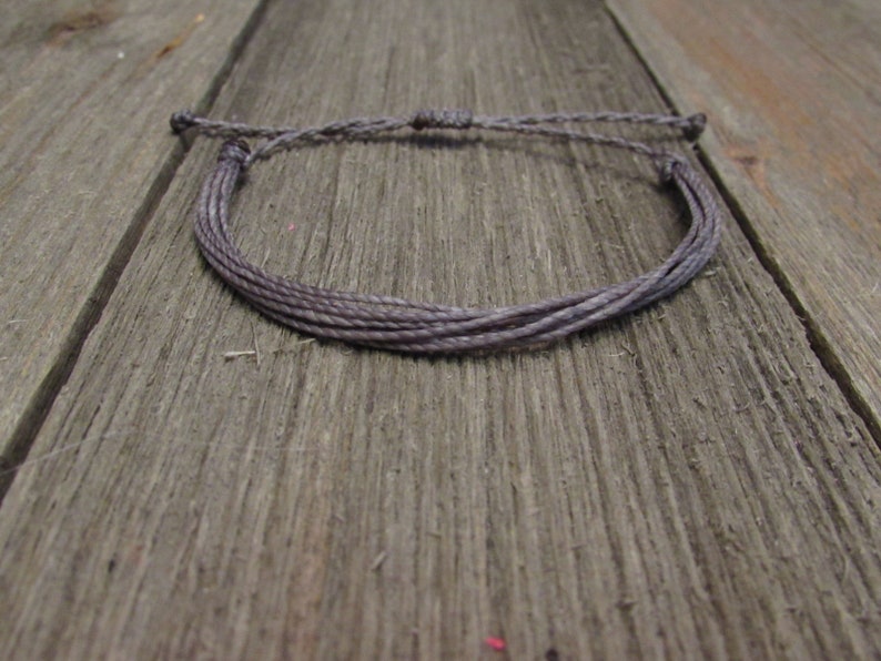 Op de afbeelding: Een grijs gevlochten armband gemaakt van meerdere strengen koord. De armband is vastgemaakt met een simpele knoop en ligt op een houten oppervlak.