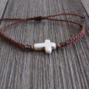 May include: A brown braided cord bracelet with a white cross charm and brown beads.