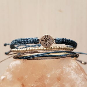 May include: A set of four bracelets with a compass charm. The bracelets are made of blue, white, and gray cord. The compass charm is silver and has the letters N, S, E, and W engraved on it.