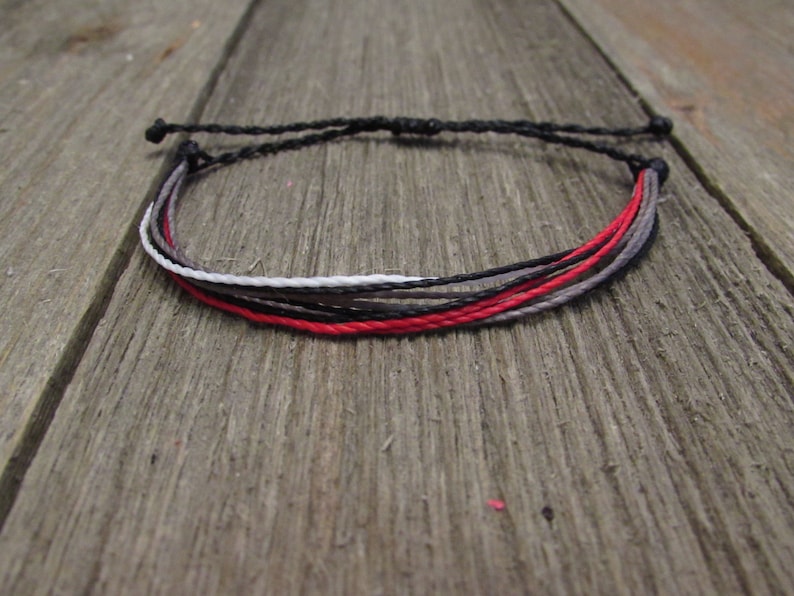 May include: A multi-strand bracelet featuring a combination of black, red, gray, and white cords. The bracelet is adjustable with a sliding knot closure. The cords are tightly woven together, creating a simple yet stylish accessory.